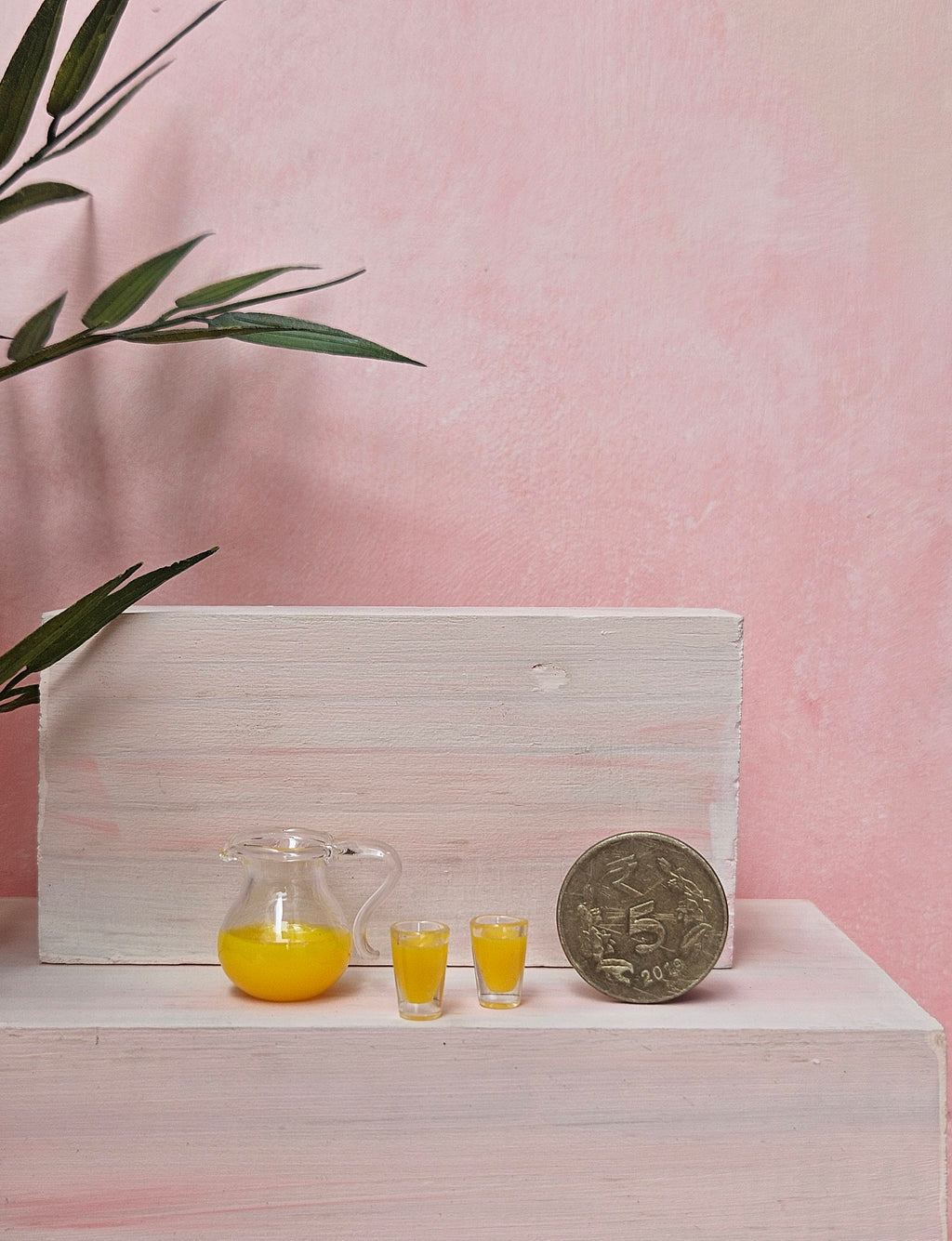 Small pitcher and glasses of yellow liquid with a coin on a white surface against a pink background