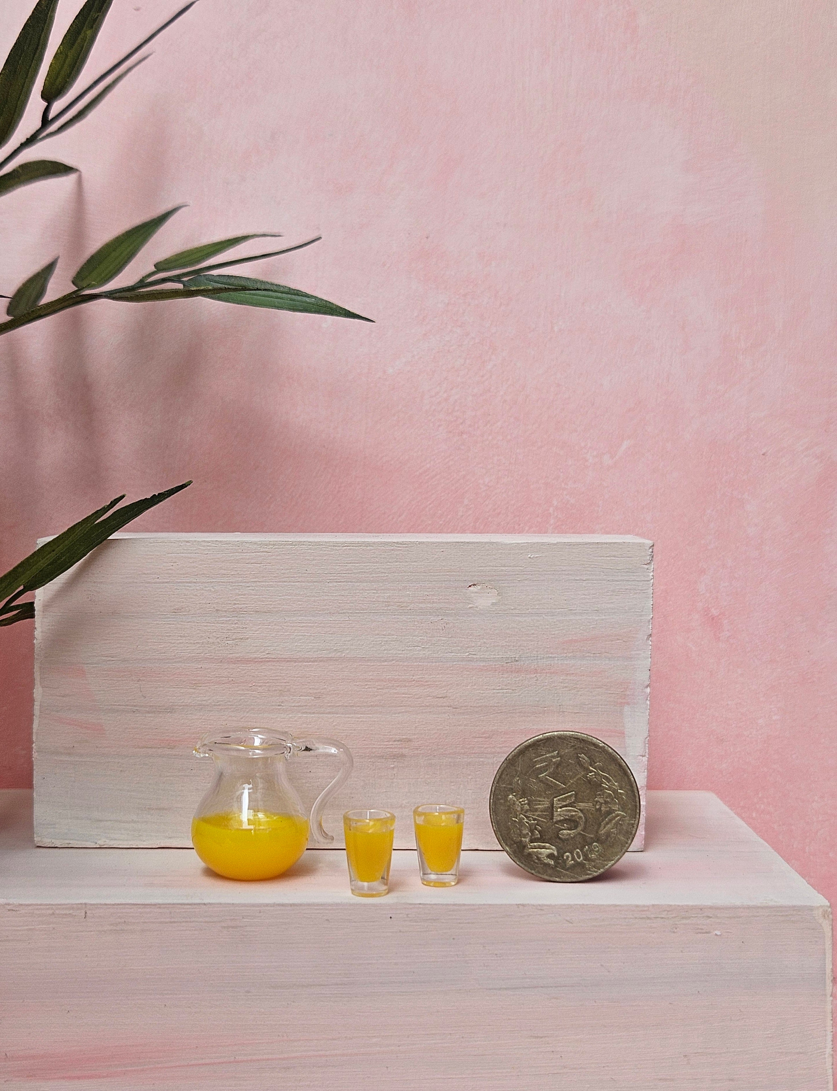 Small pitcher and glasses of yellow liquid with a coin on a white surface against a pink background