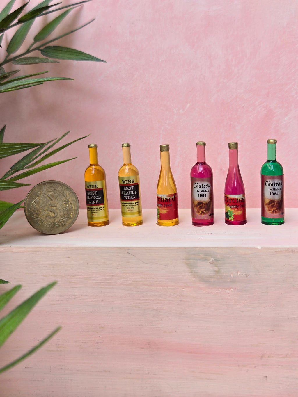 Set of six miniature wine bottles on a pink surface with a plant in the corner.