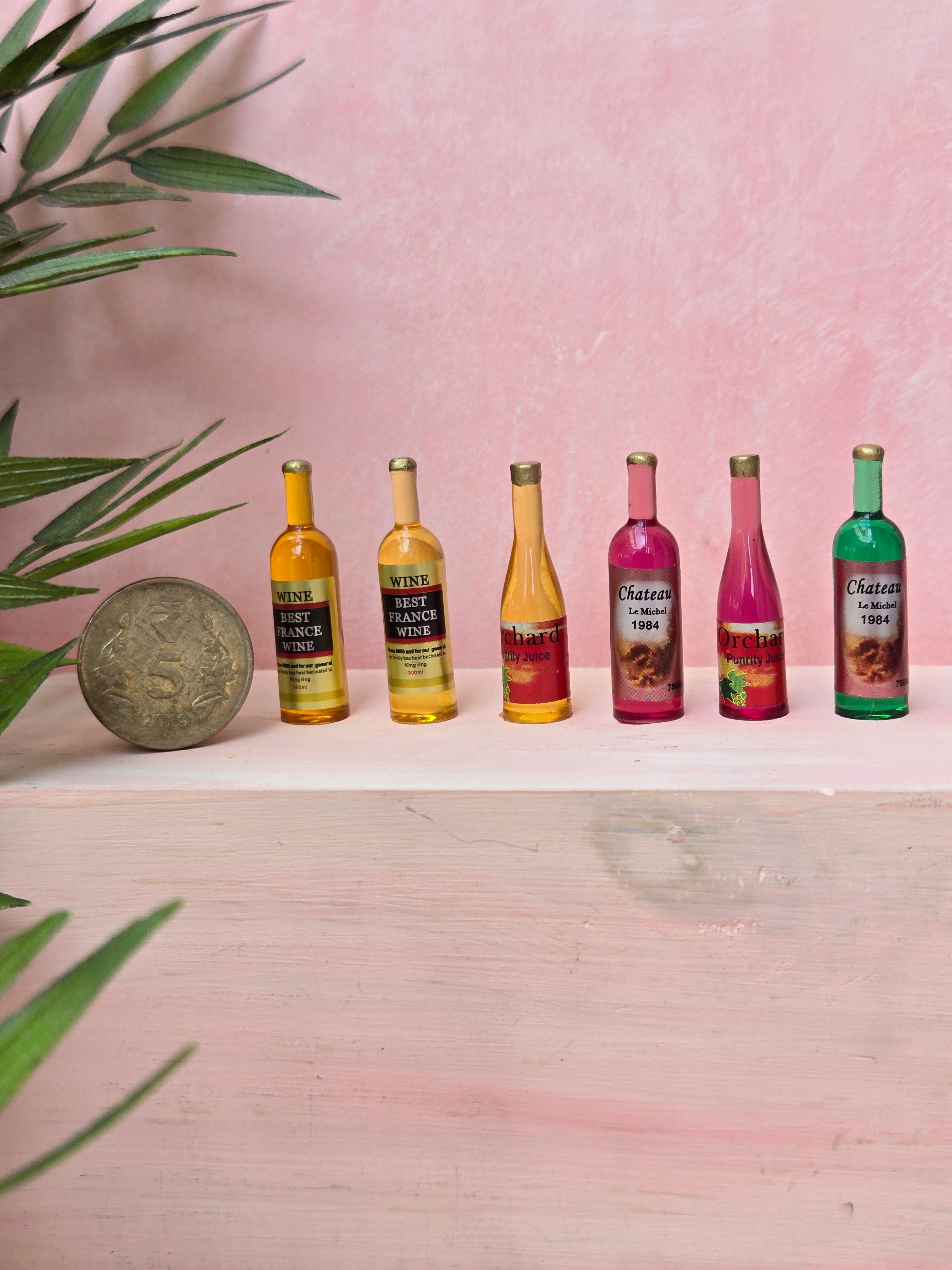 Set of six miniature wine bottles on a pink surface with a plant in the corner.