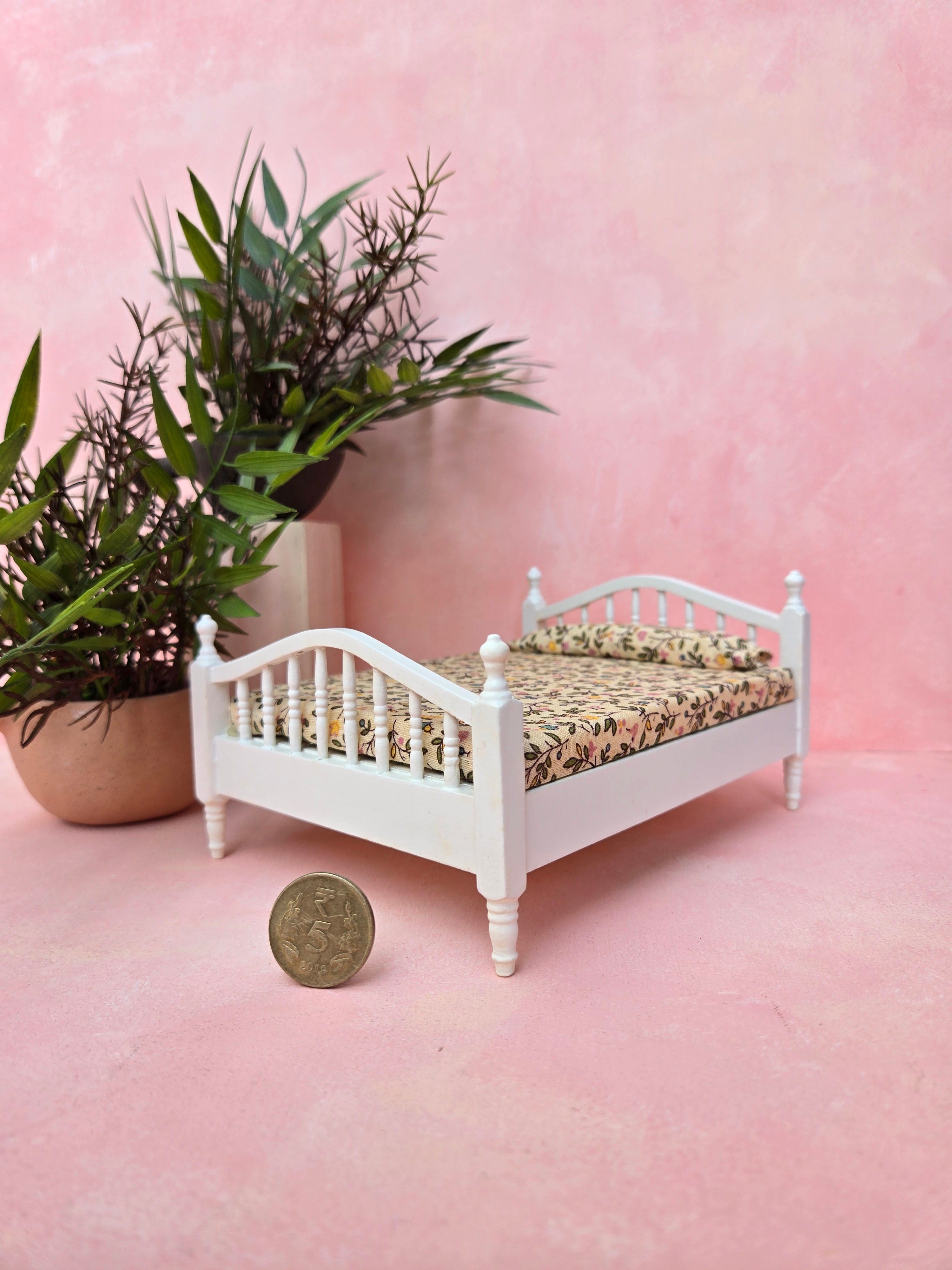 Miniature white bed with floral bedding on a pink surface, next to a potted plant.