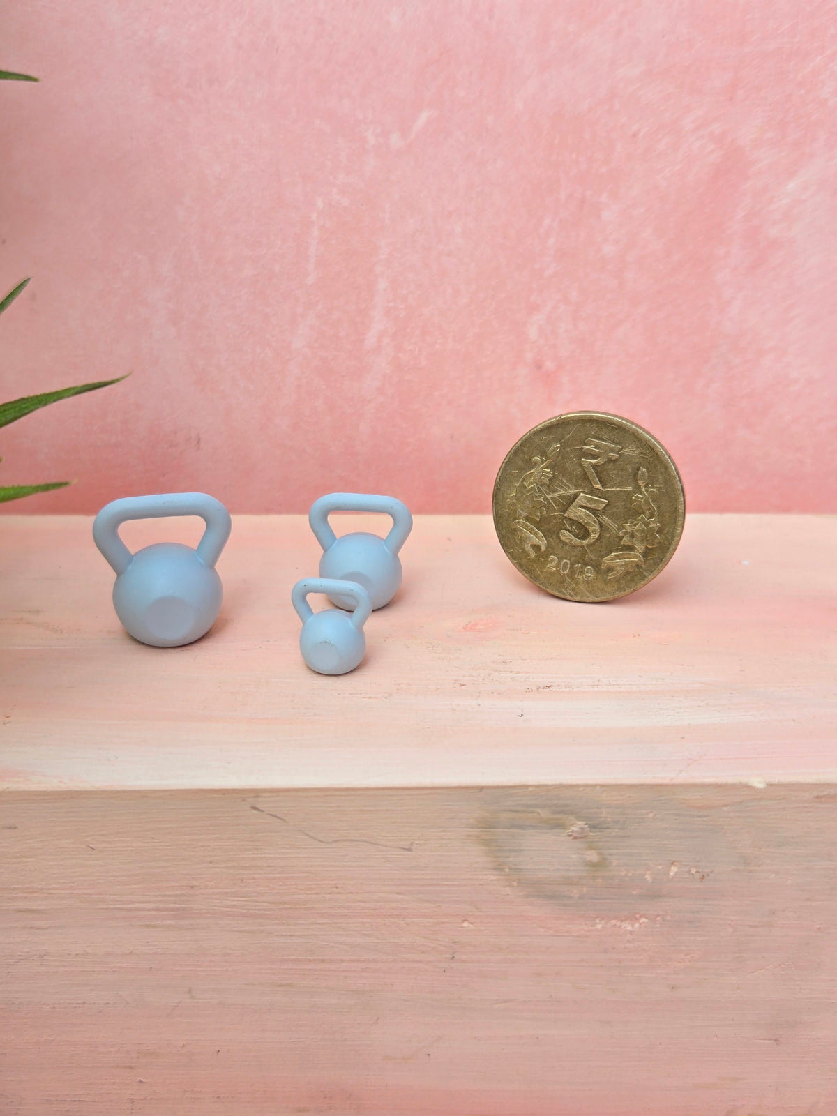 Two small blue dumbbells next to a coin on a wooden surface with a pink background