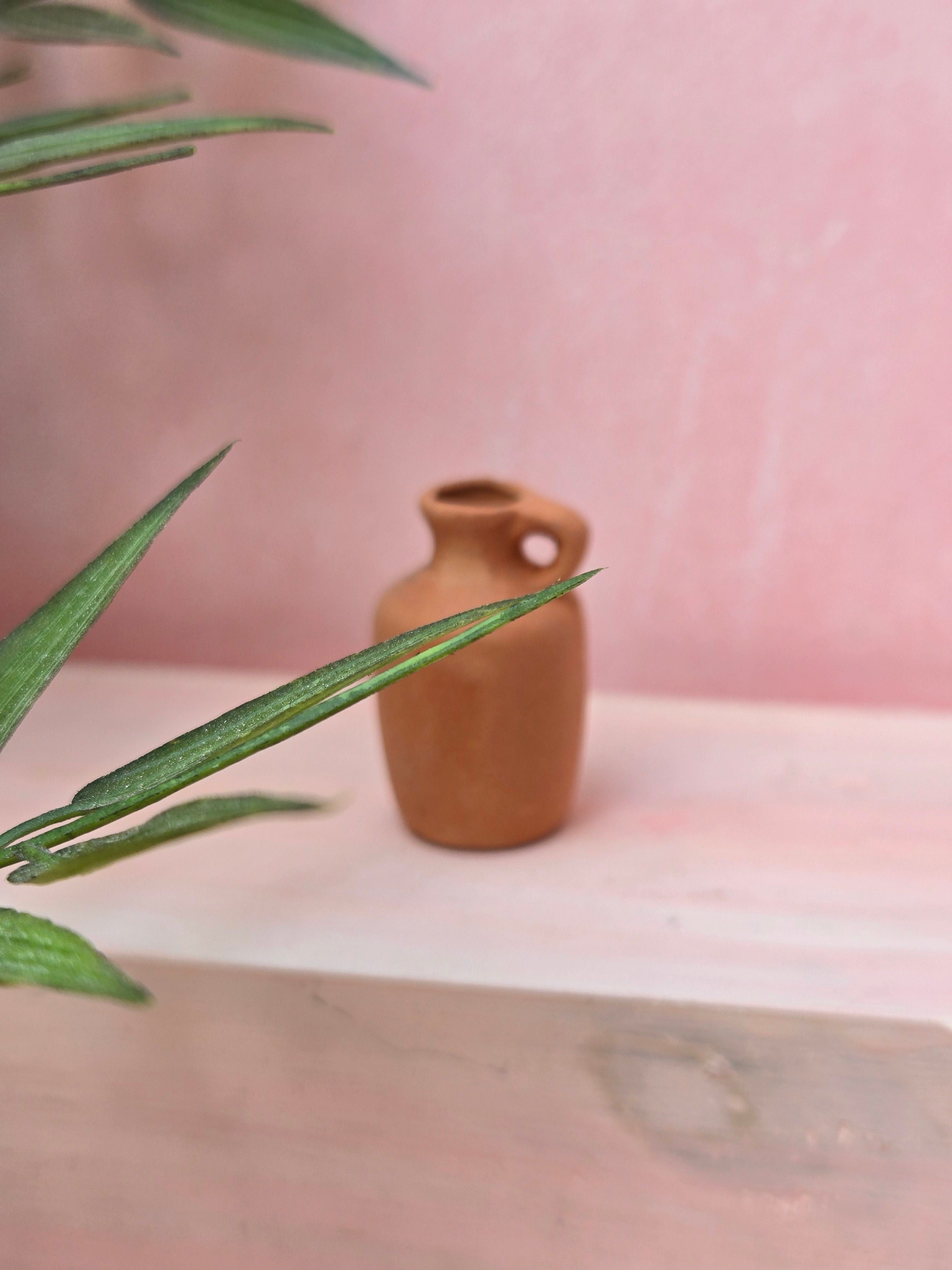 Small brown ceramic vase on a light pink surface with green leaves in the foreground.