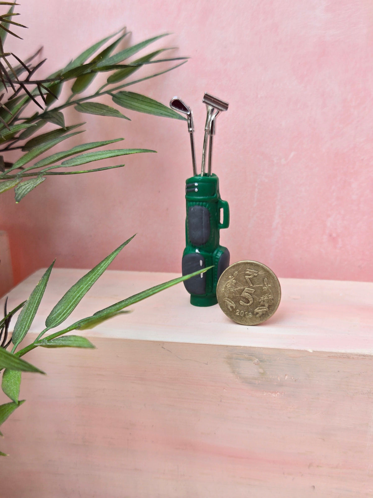 Green golf club set in a miniature golf bag with a coin for scale on a pink background