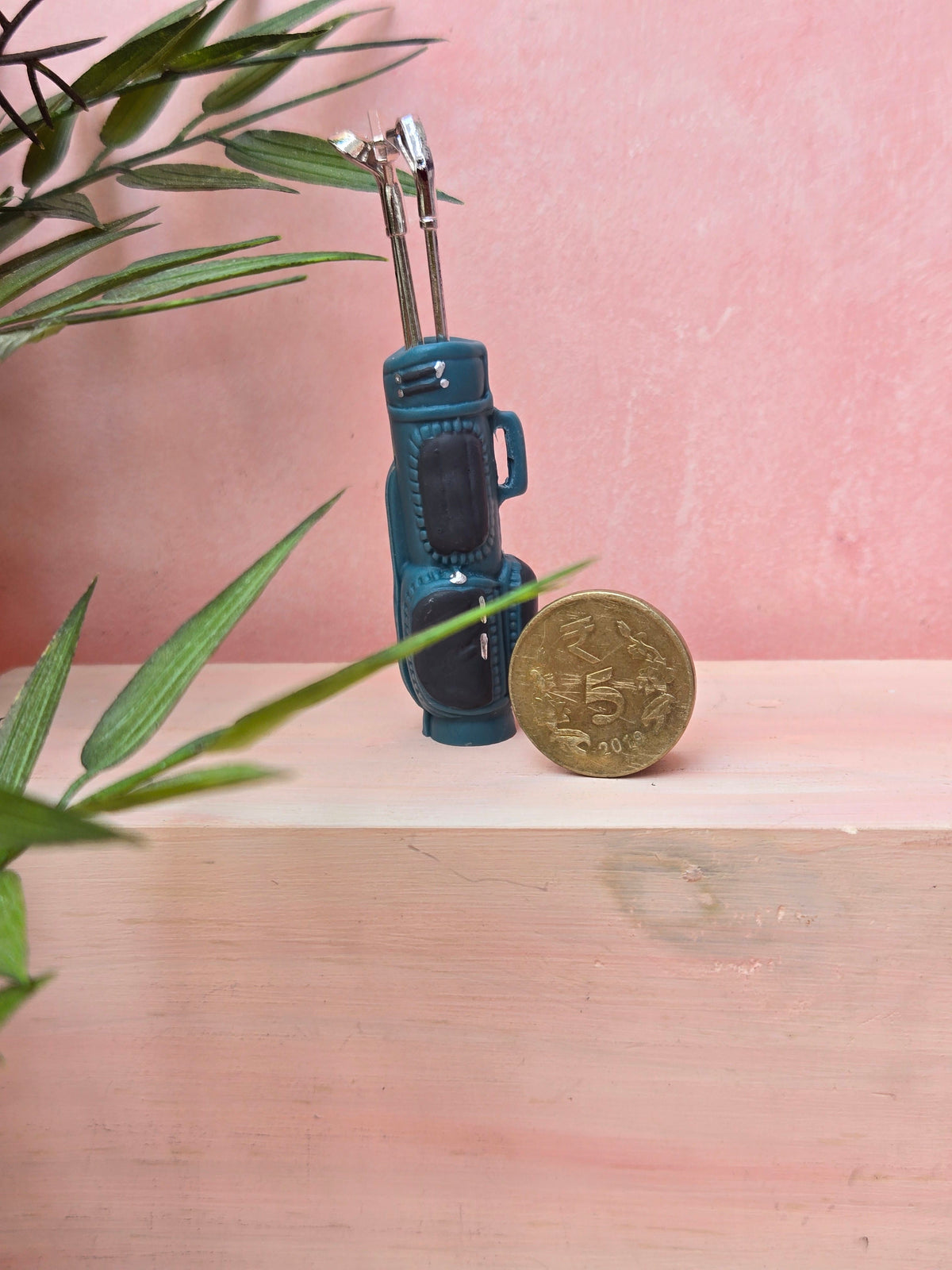 Miniature golf bag next to a coin on a wooden surface with a pink background