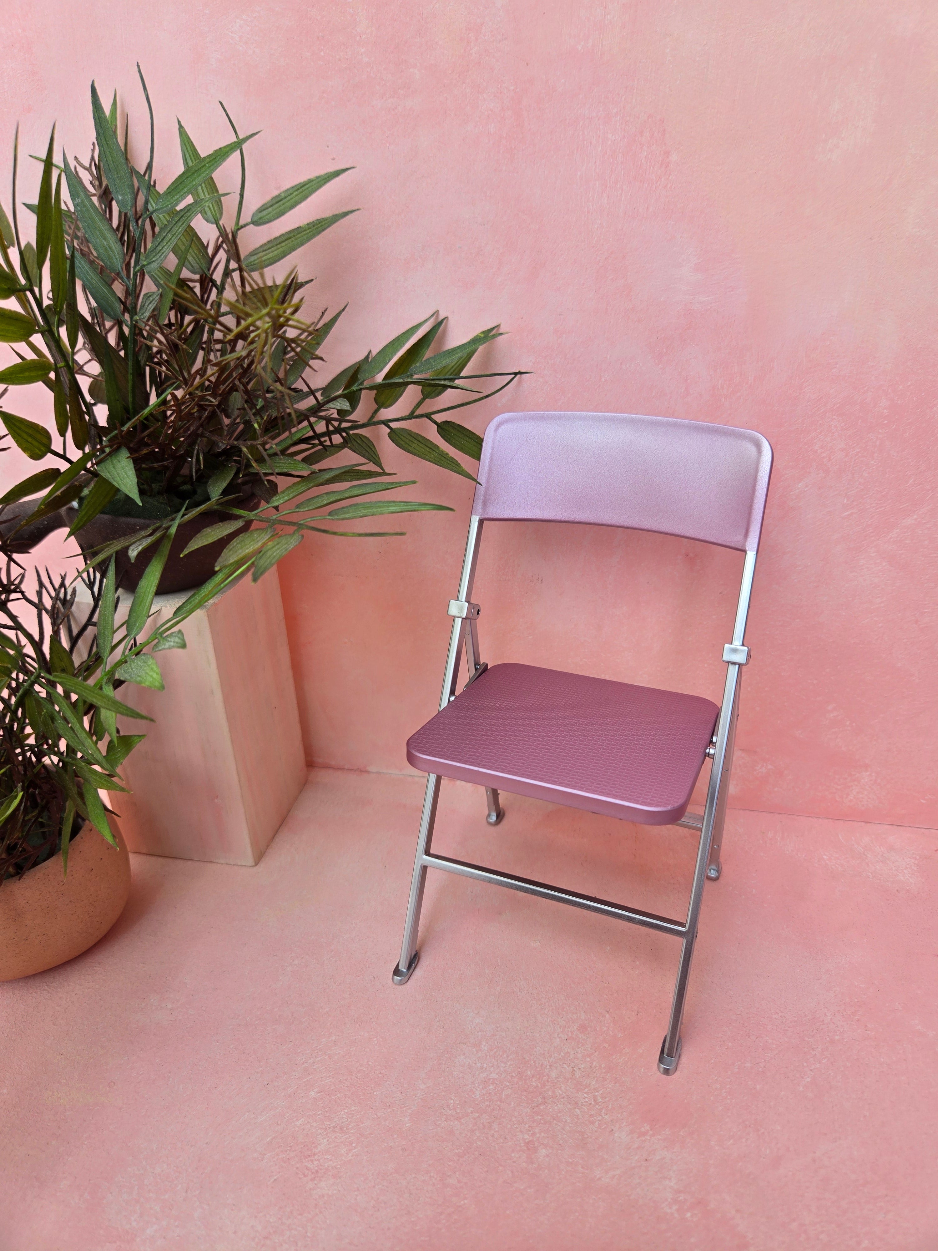 Pink folding chair against a pink wall with a plant to the left.