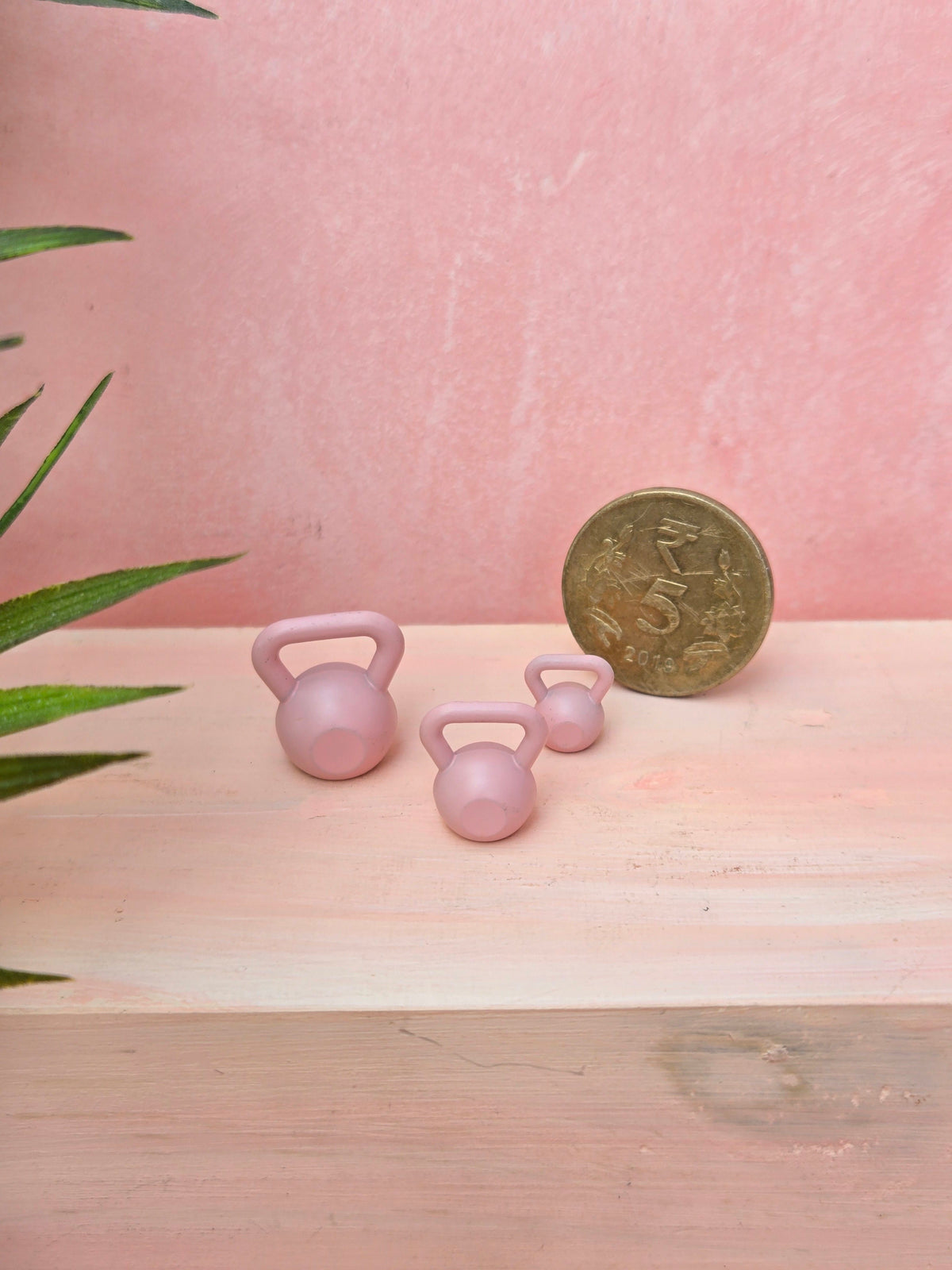 Two small pink kettlebells next to a gold coin on a wooden surface with a pink background.
