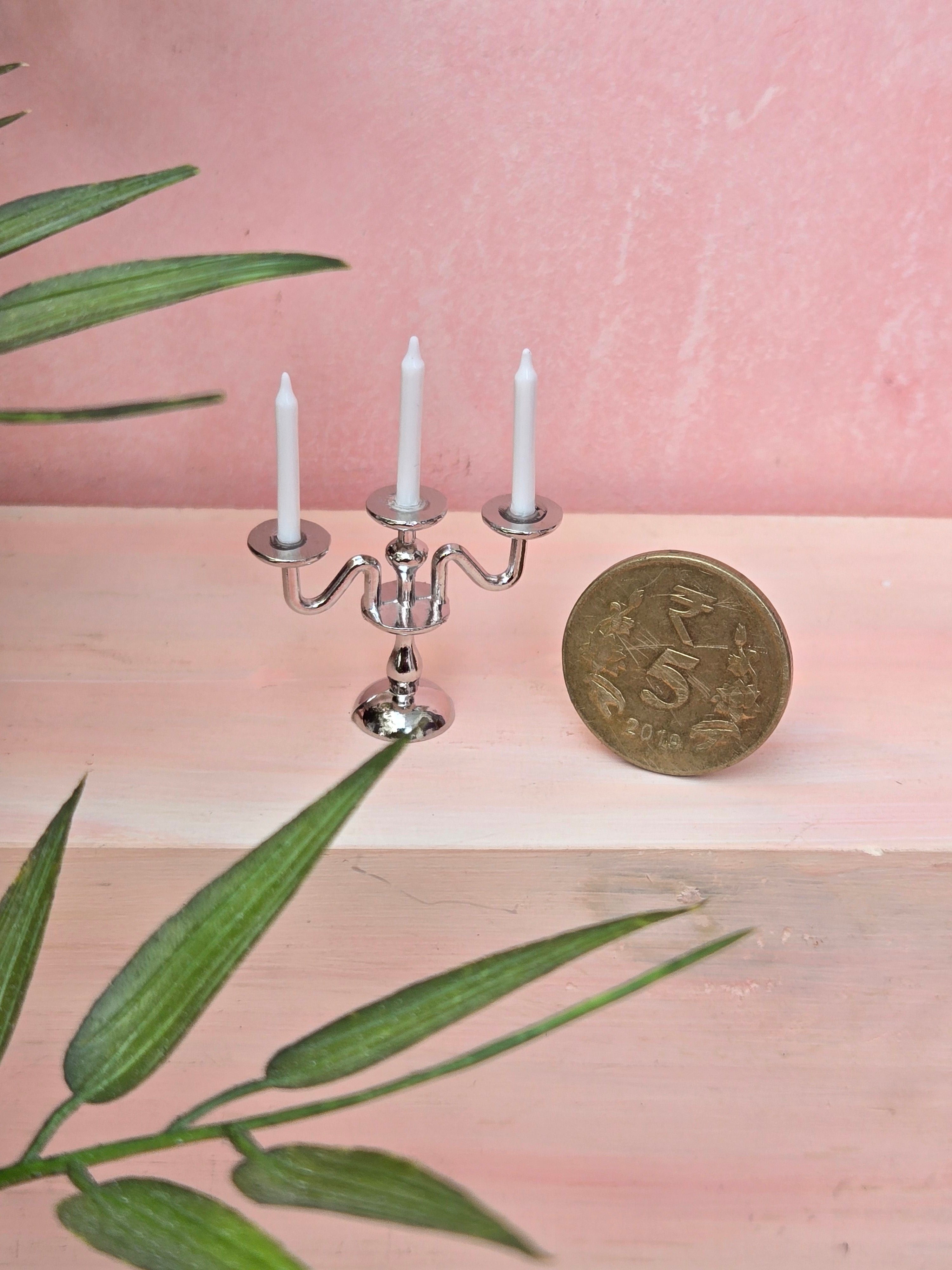 Small silver candelabra with three white candles on a pink surface, accompanied by a gold coin and green leaves.