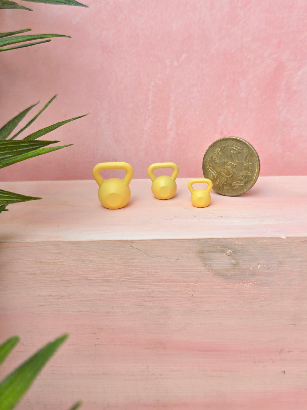 Three yellow miniature kettlebells and a coin on a pink surface with green leaves.